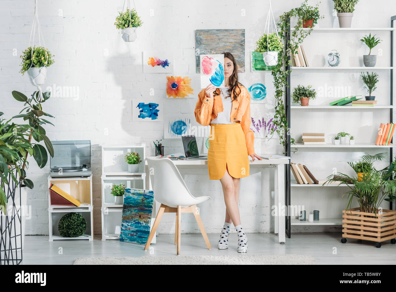 trendy young woman covering face with painting while standing in spacious room decorated with green plants and drawings on white wall Stock Photo