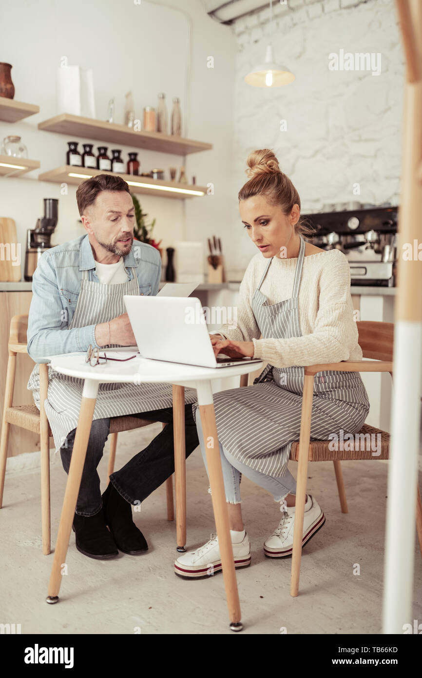 Working side by side. Two focused business partners sitting in front of kitchen equipment in their cafe and working. Stock Photo