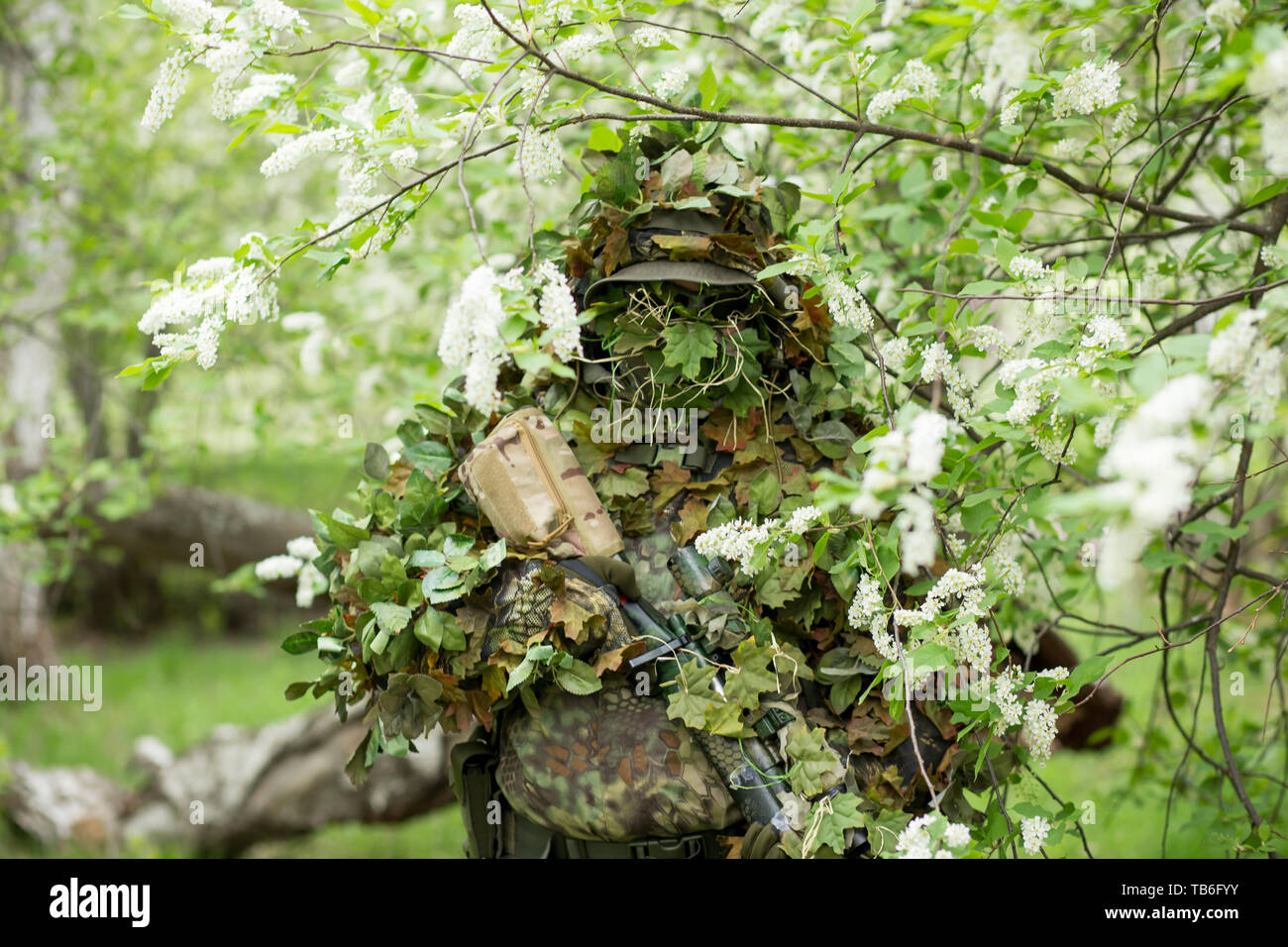 Cute portrait of a military male sniper in camouflage, mask, cap and rifle on his hands against the background of a flowering cherry in the summer out Stock Photo