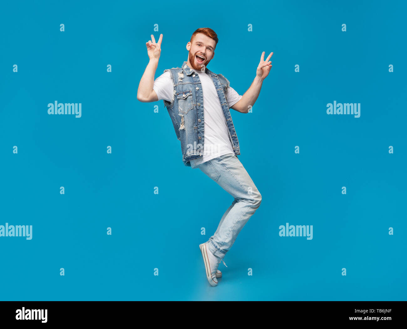 Awesome guy showing peace gesture and standing on tiptoes on blue background, copy space Stock Photo