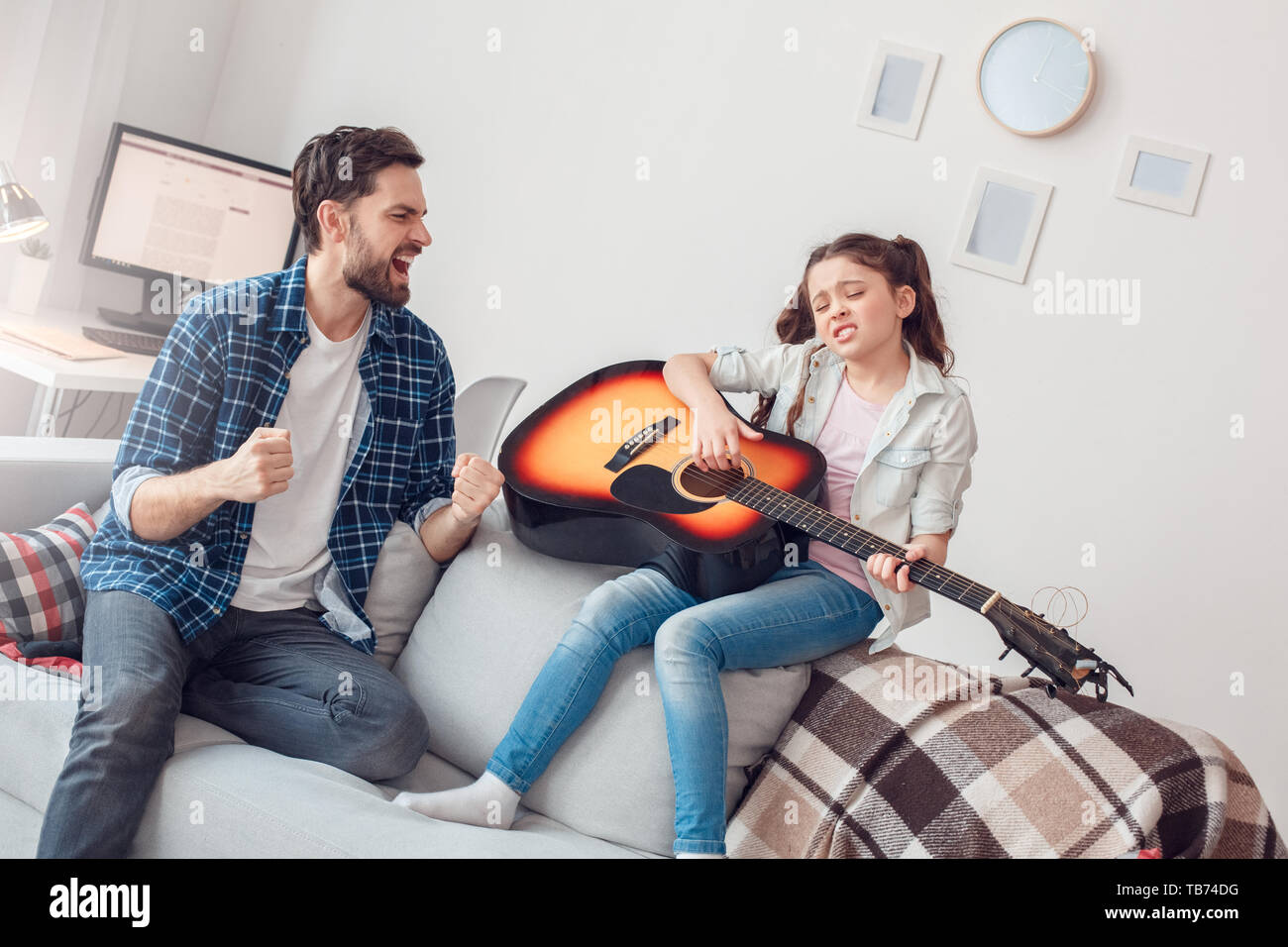 Bearded man and little girl at home family time sitting on sofa daughter playing guitar singing with father smiling joyful Stock Photo
