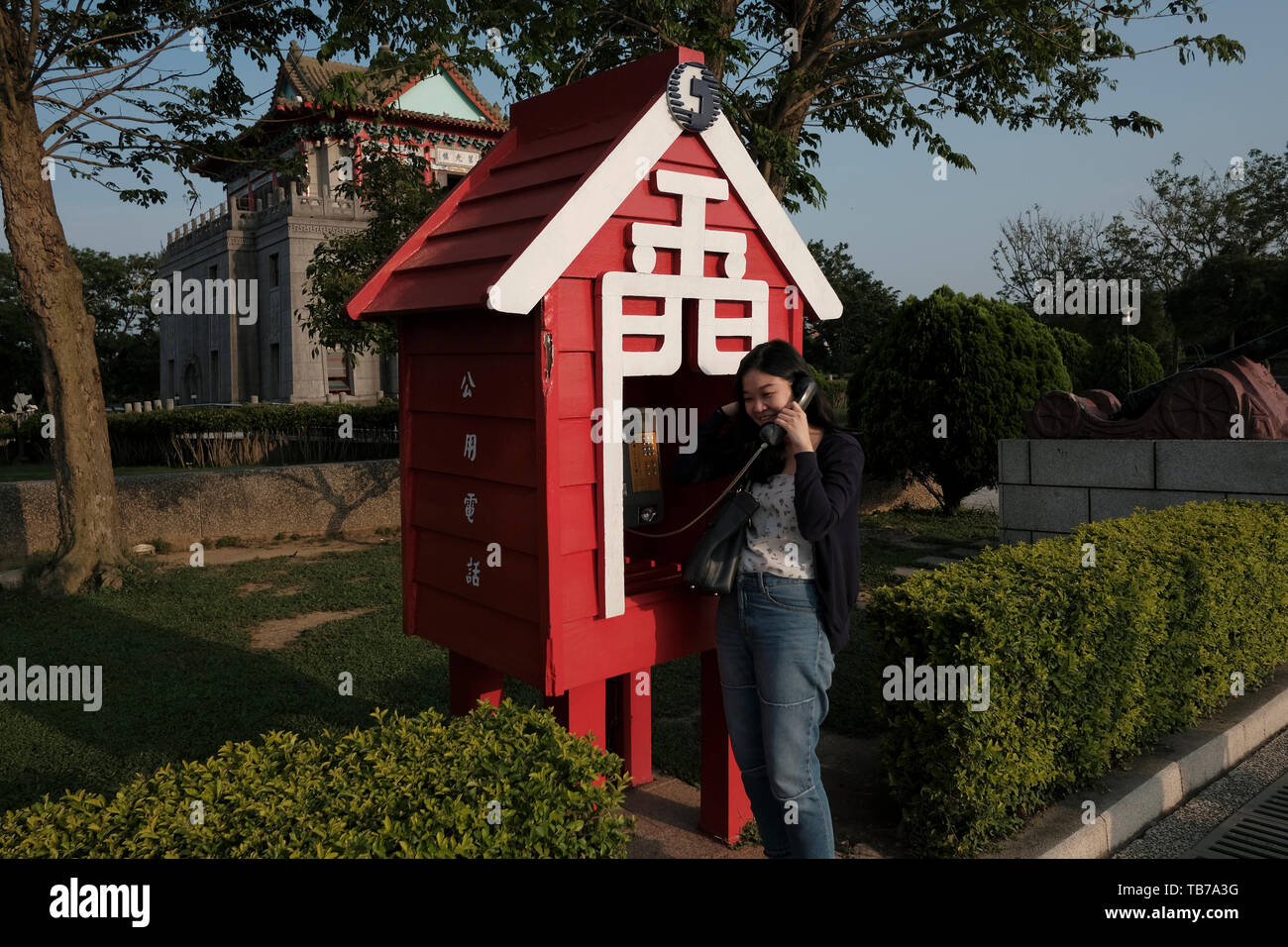 Woman speaking in a red phone booth with the word Jīnmen or Golden Gate which means Kinmen in front of Ju Guang Lou in Jincheng Township, Kinmen County or island, Taiwan Stock Photo