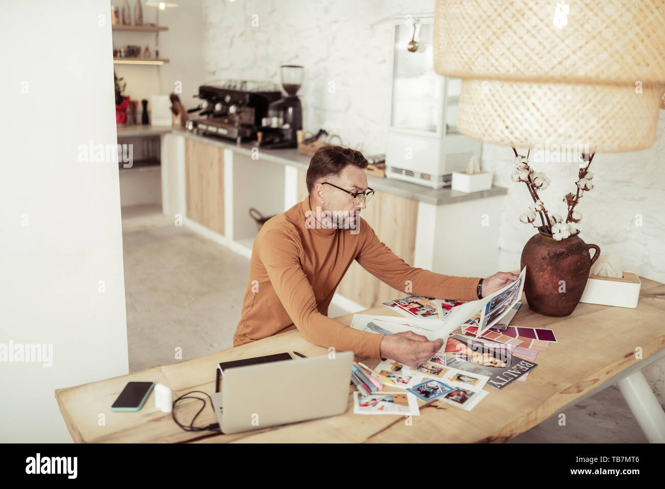 Analysing work. Serious fashion designer sitting at the table looking at his sketches and pictures of dresses comparing them. Stock Photo