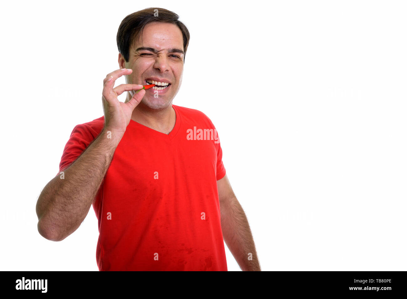 Studio shot of young happy Persian man smiling while biting red chili ...