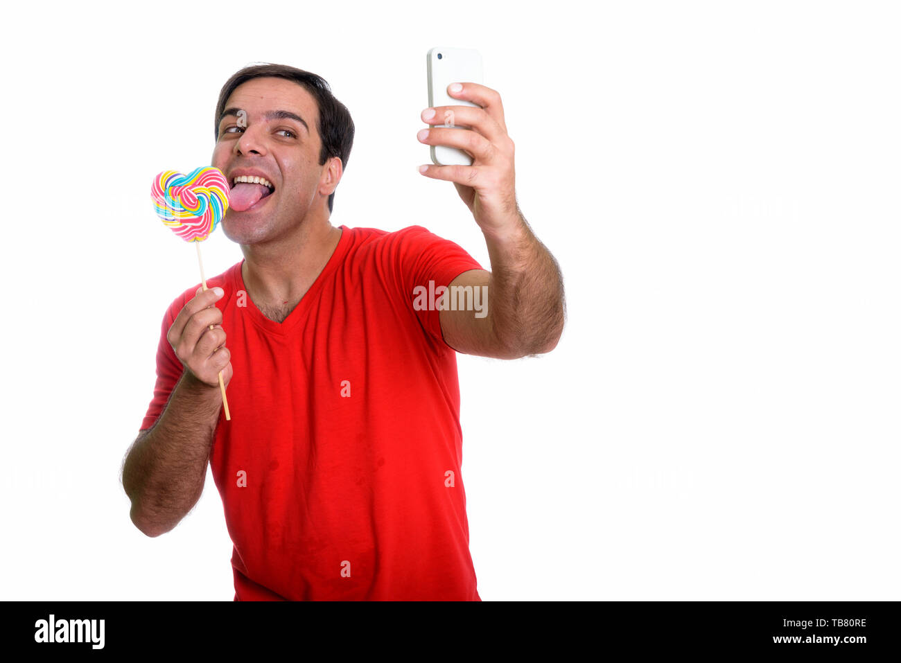 Studio shot of young happy Persian man smiling and taking selfie picture with mobile phone while licking heart shaped lollipop isolated against white  Stock Photo