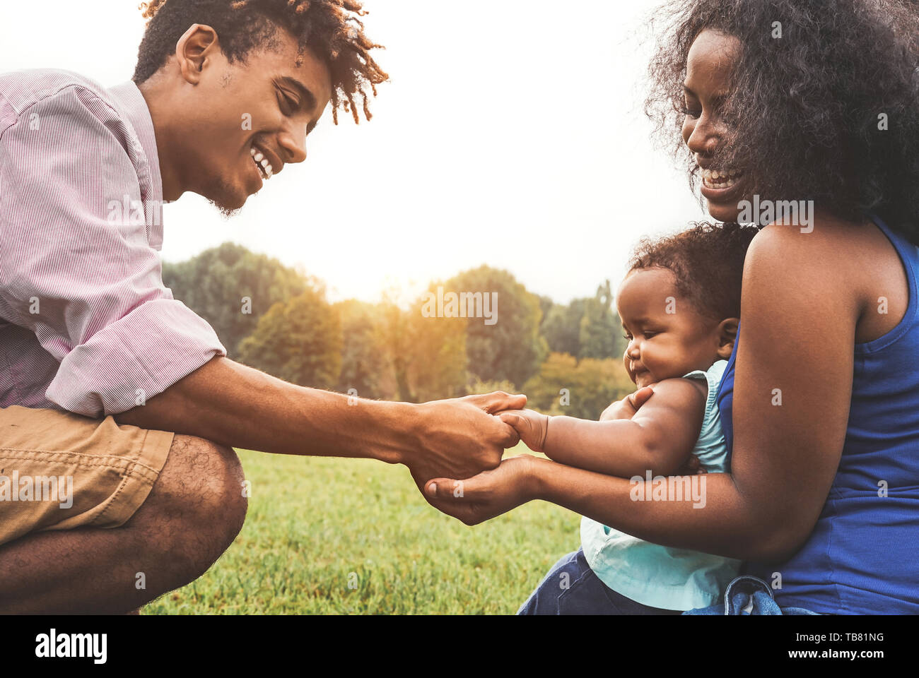 Happy African family enjoying time together in public park - Mother and father having fun playing with their daughter at sunset outdoor Stock Photo