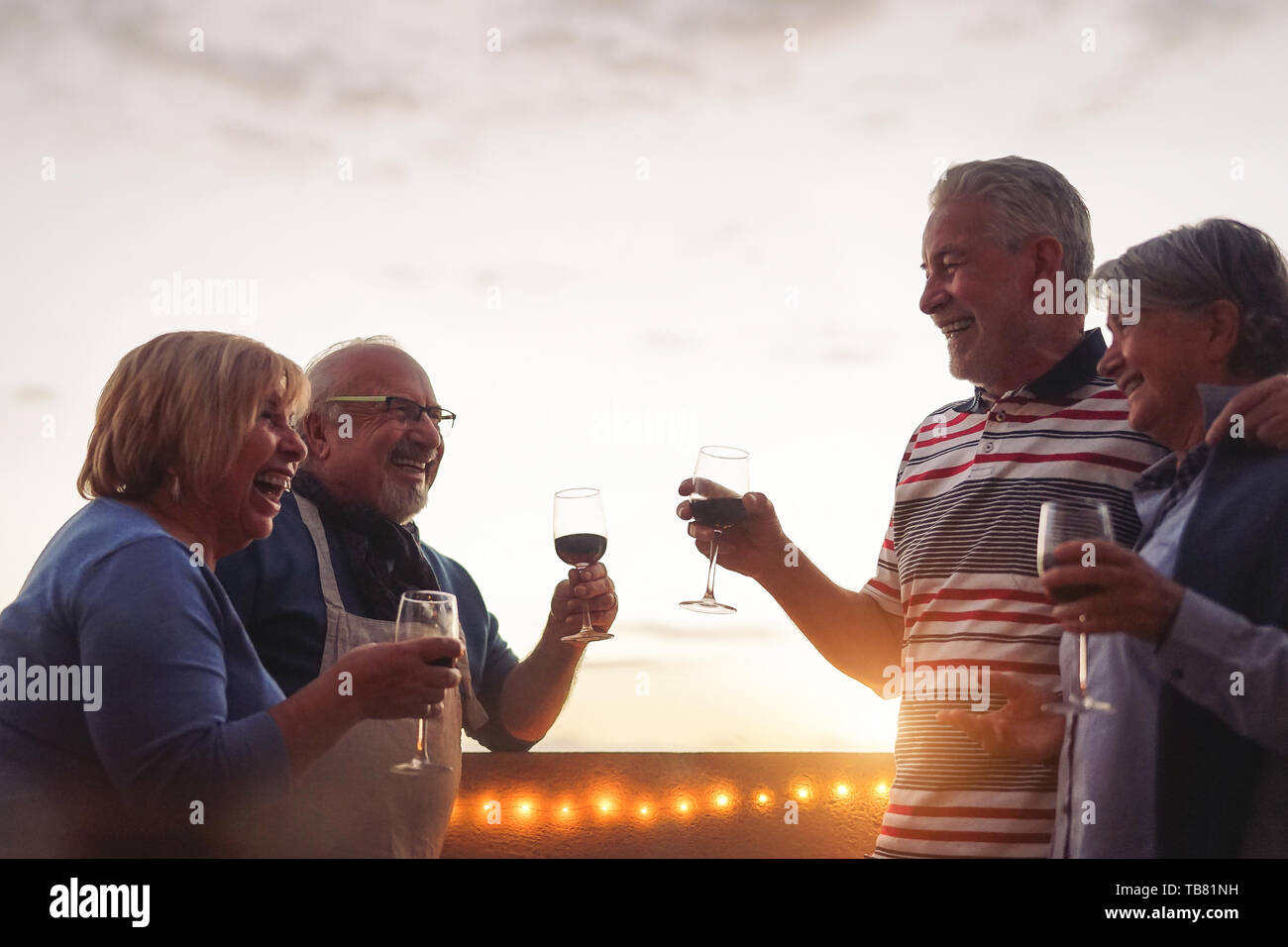 Happy senior friends drinking red wine at barbecue dinner in terrace - Mature people cheering and laughing together at sunset on rooftop Stock Photo