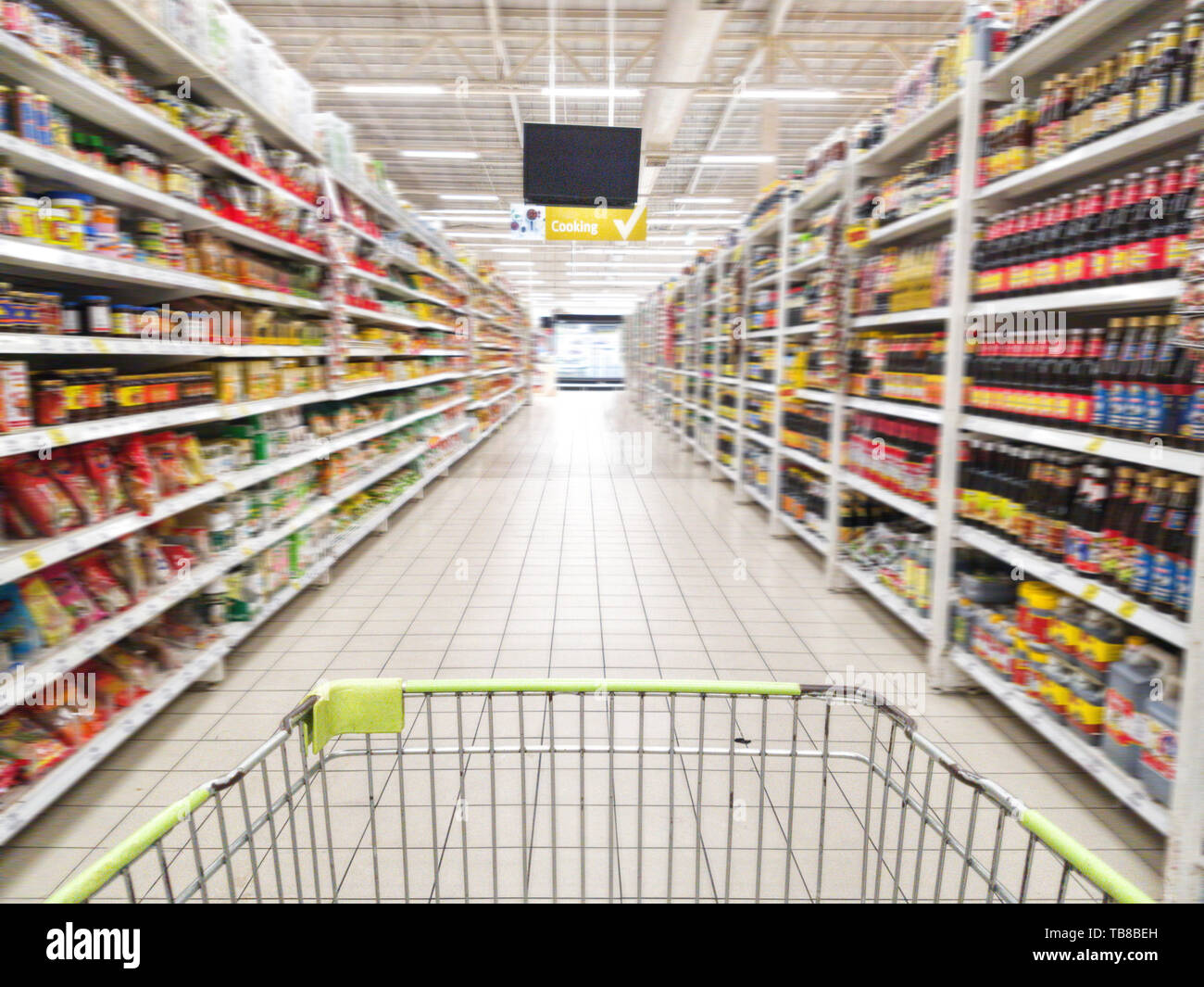 shopping cart with abstract blurred supermarket aisle with colorful goods on shelves at background, sign of product for cooking hanging from ceiling Stock Photo