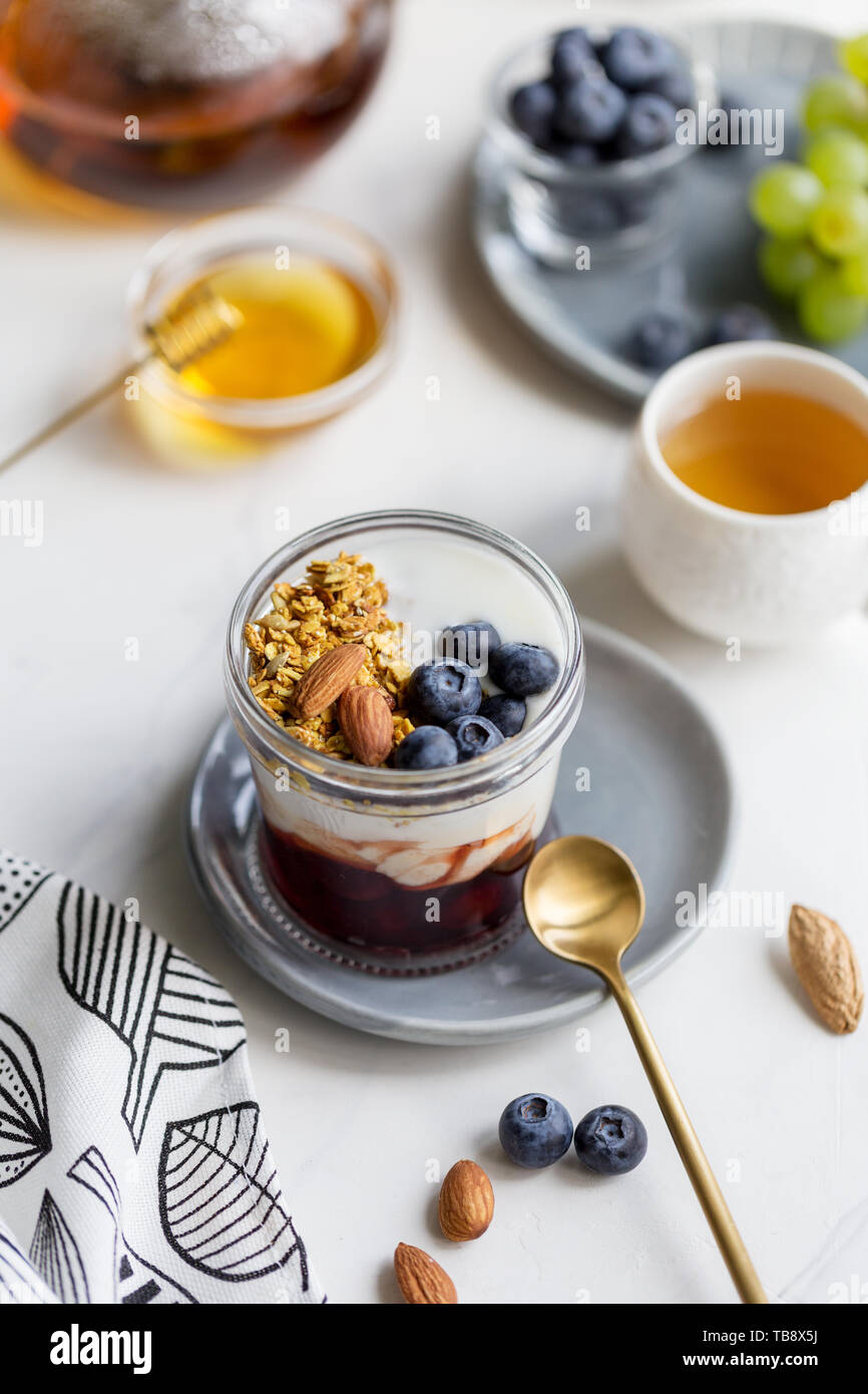 Tasty cereal with blueberries, almond, jam and yogurt in jar on bright background. Honey, tea, grape, napkin and spoon around. Concept of healthy brea Stock Photo