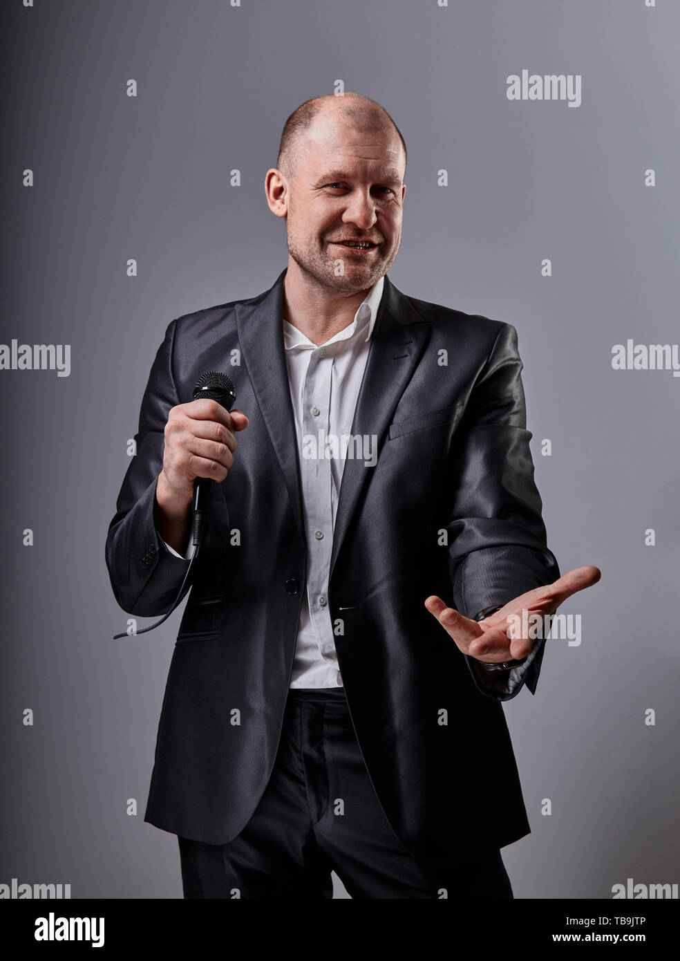 Fun emotional moving happy performer man presenting the show holding microphone in hand and showing the hand the welcome sign on grey color background Stock Photo