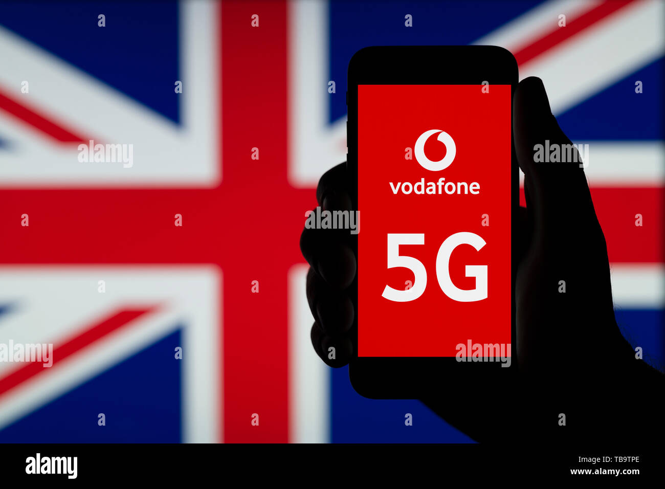 A silhouetted hand of a man holds a smartphone displaying the logo of Vodafone and the letters 5G, infront of a UK flag (Editorial use only). Stock Photo