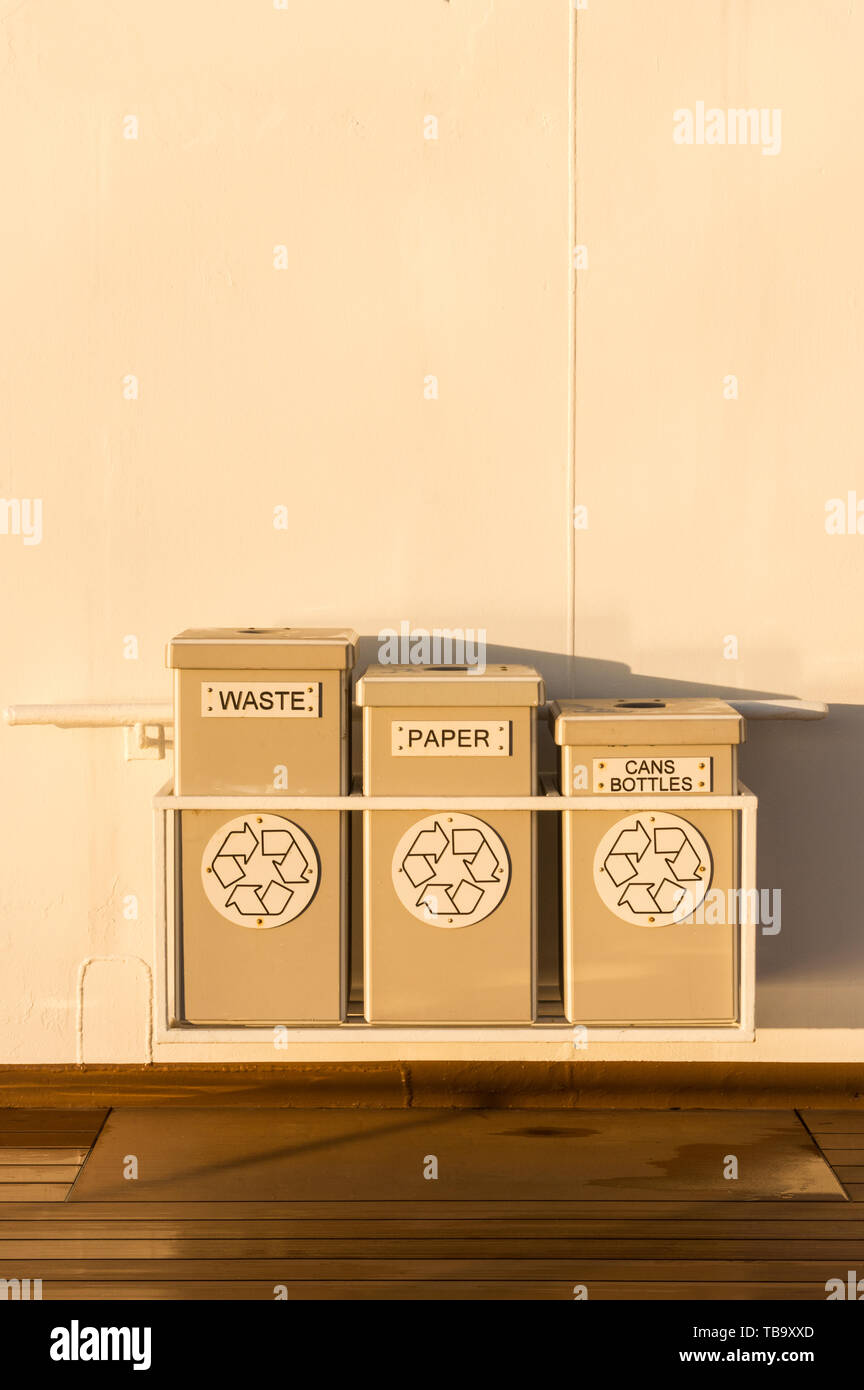 Three tidy waste and recycling receptacles hung on painted white metal exterior wall of cruise ship. Stock Photo