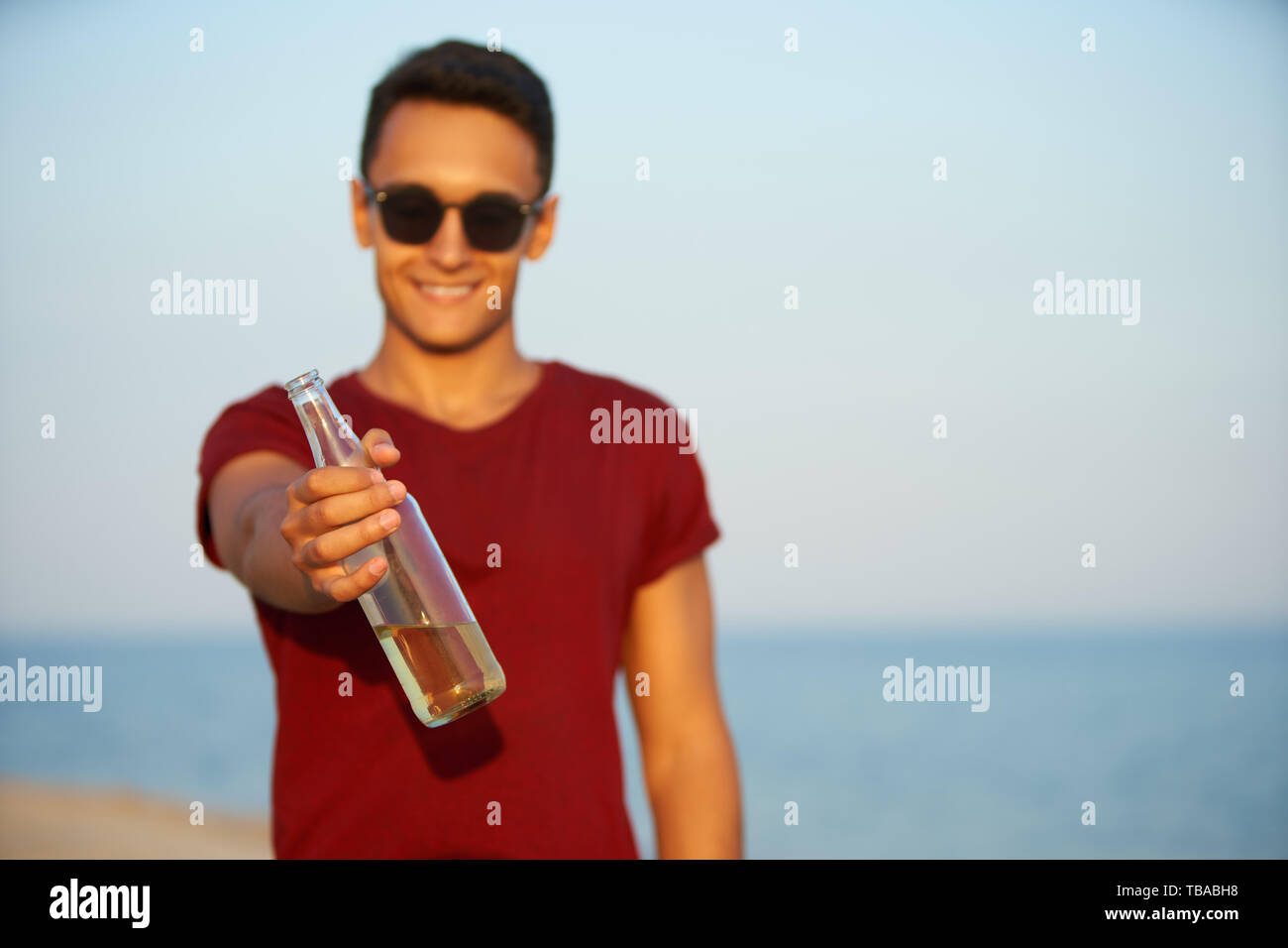 Young handsome man drinking beer on the beach during sunset. Male quenches thirst with lemonade beverage at sandy sea shore. Guy proposes or offers Stock Photo