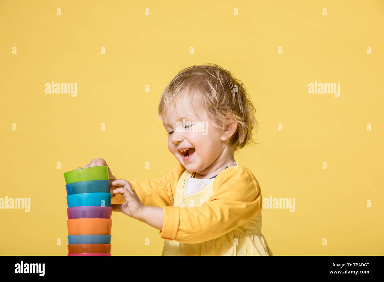 Young Blonde Girl in Yellow Dress Playing with Colorful Cups and Cheerfully laughing on Yellow Background Stock Photo