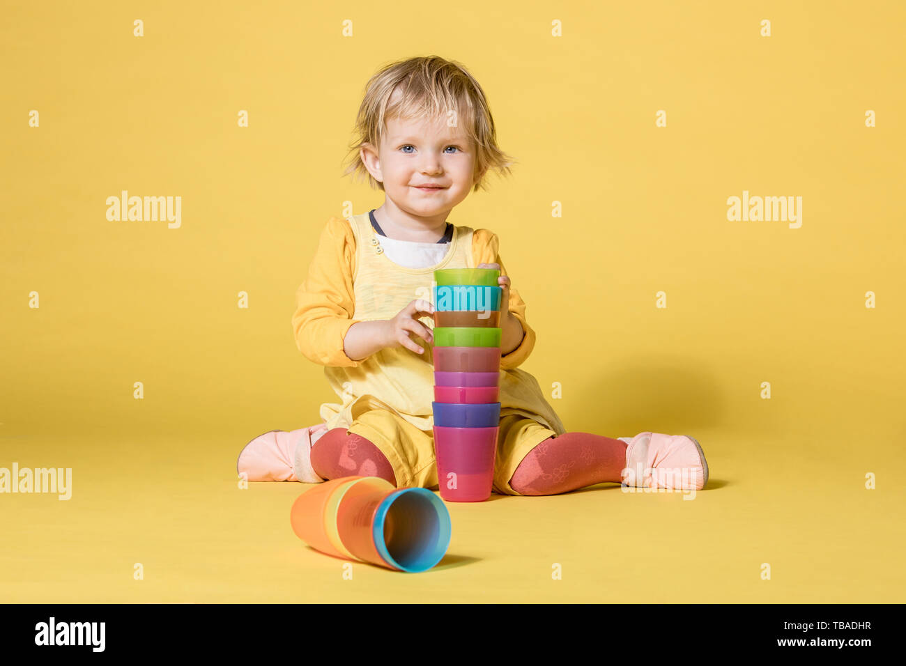 Young Blonde Girl in Yellow Dress Playing with Colorful Cups and Cheerfully laughing on Yellow Background Stock Photo