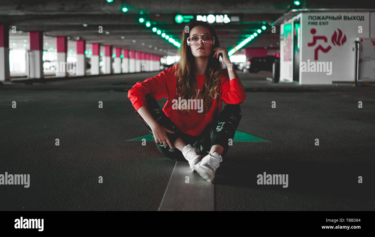 Pretty young woman in red hoodie sitting on car parking. Wearing stylish urban outfit Stock Photo