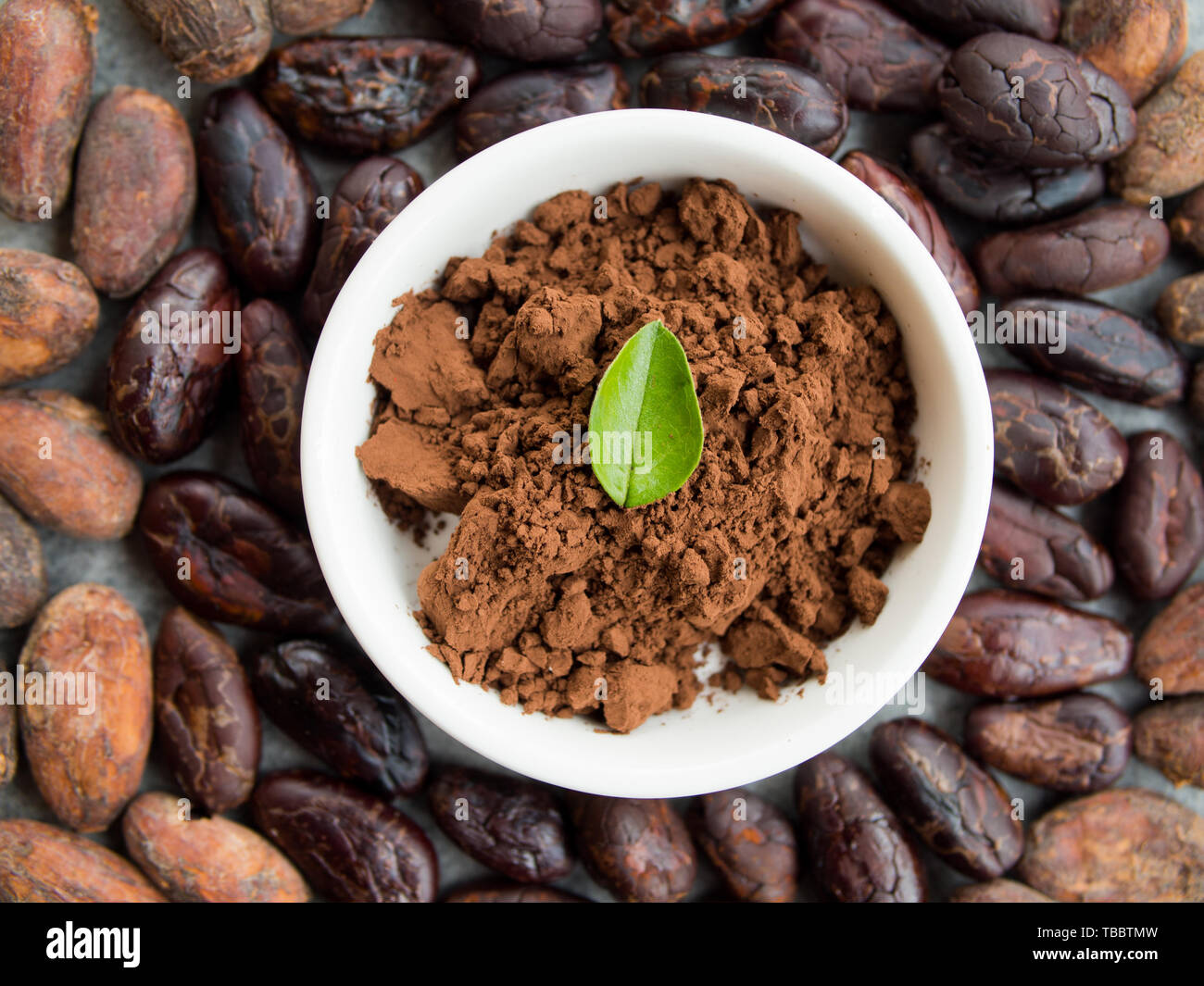 Cocoa powder in a small bowl, roasted cocoa beans around, top view. Stock Photo