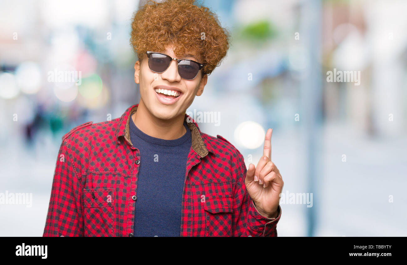 Young handsome man with afro hair wearing sunglasses pointing finger up ...