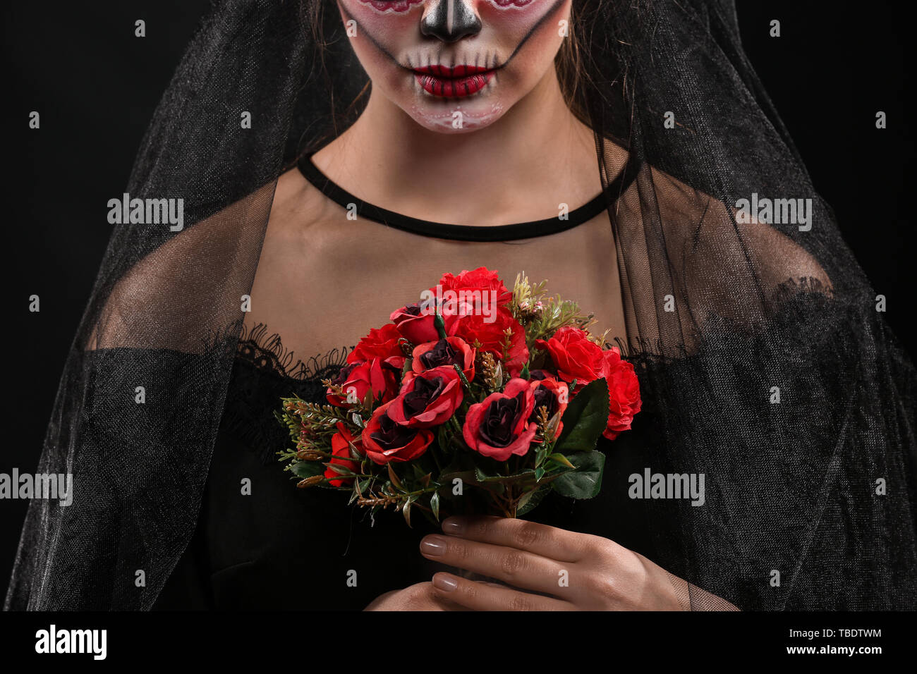Young woman with painted skull on her face for Mexico's Day of the Dead and flowers against dark background Stock Photo