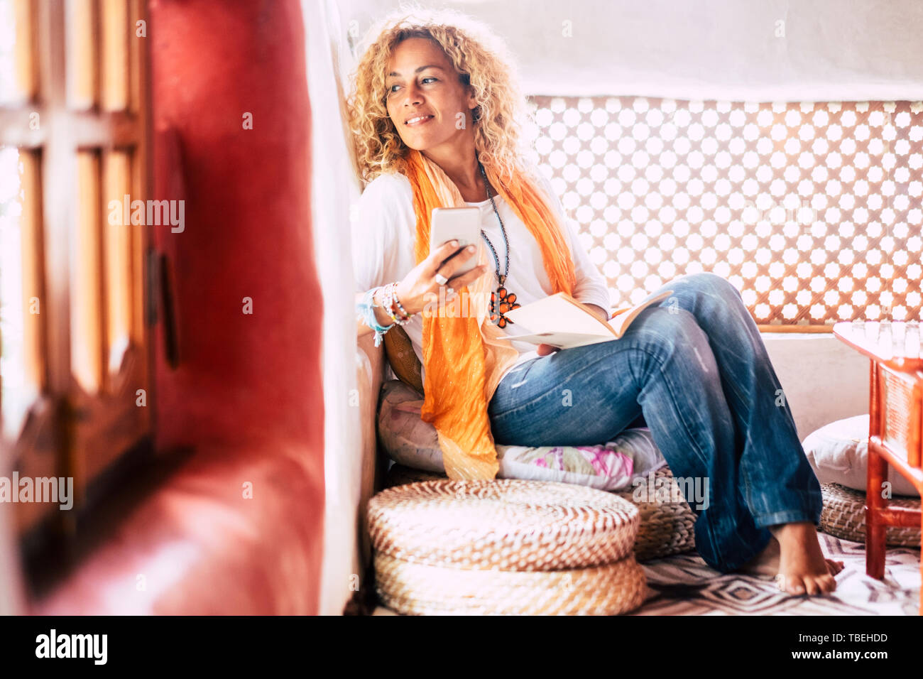 Beautiful adult young blonde woman sit down at home with paper book and mobile phone looking outside the window - relax at home and no stress lifestyl Stock Photo