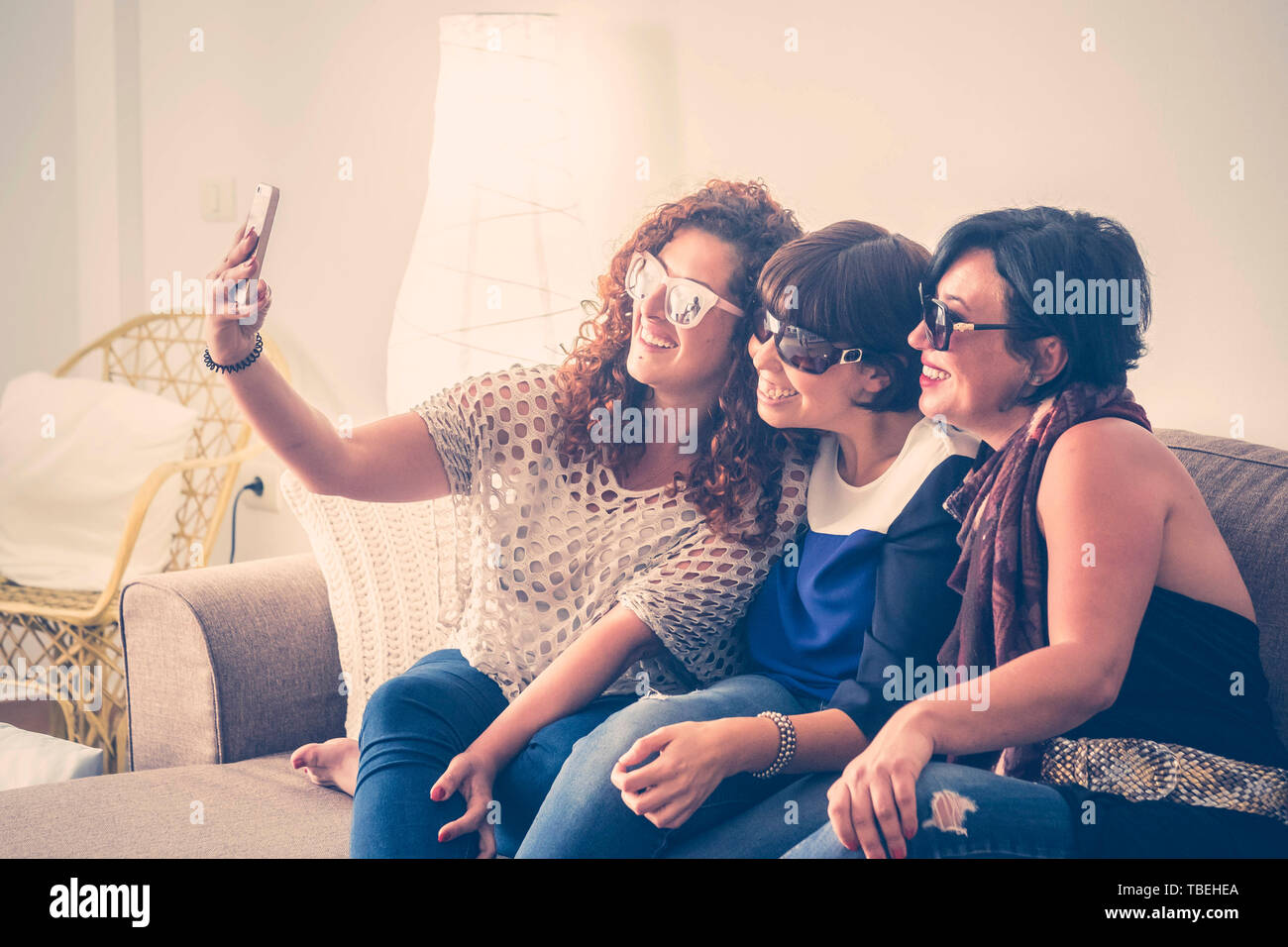 Friend group indoor caucasian young beautiful females doing a picture in selfie style with a mobile phone at home sit down on the sofa. friendship and Stock Photo