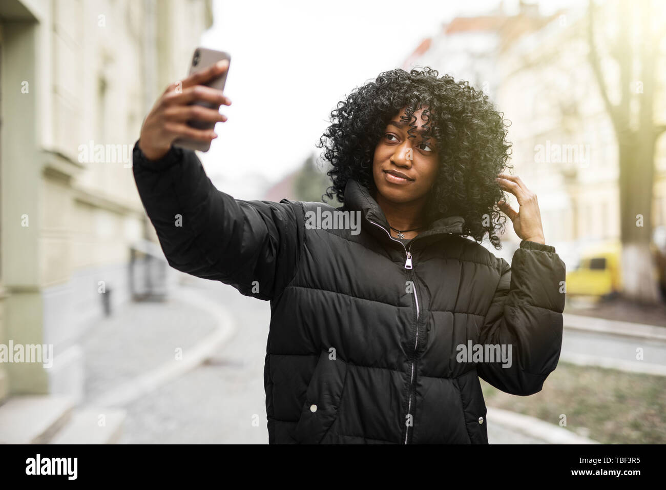 Cheerful young african woman wearing coat walking outdoors, taking a selfie Stock Photo