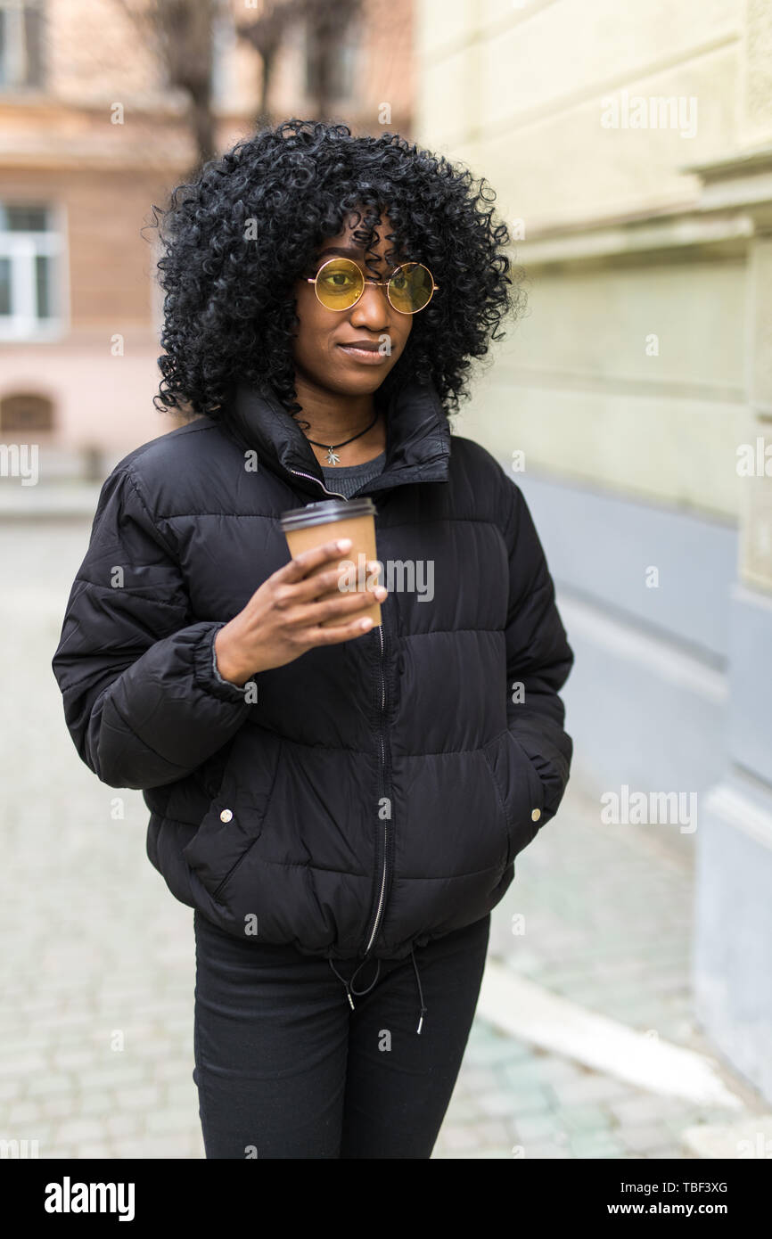 Cheerful young african woman wearing coat walking outdoors, holding takeaway coffee cup Stock Photo