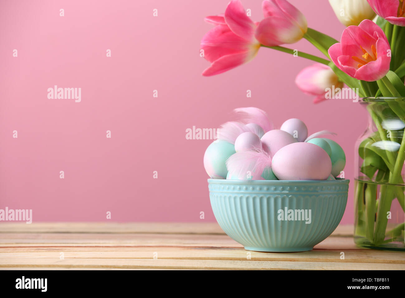 Bowl with Easter eggs and flowers on table against color background ...