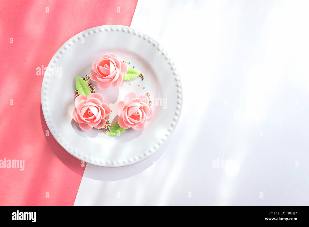 Delicate pink and white background with pastry roses and confetti on plate. Shot with mixed light. Top view, flat lay Stock Photo