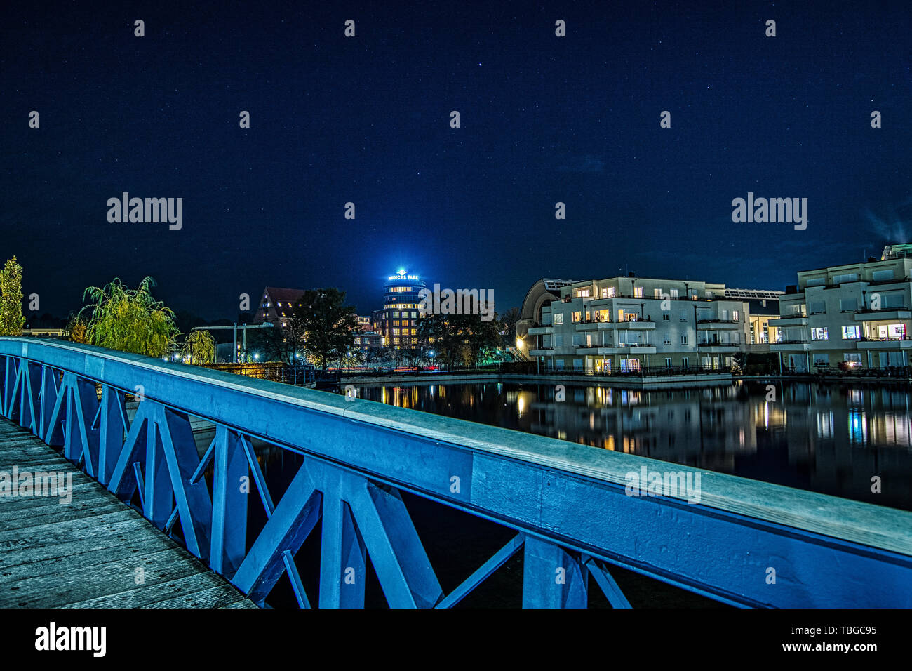 nightshot of the wooden blue bridge crossing the water of Humboldt harbour in Berlin Tegel with illuminated buildings in the background Stock Photo