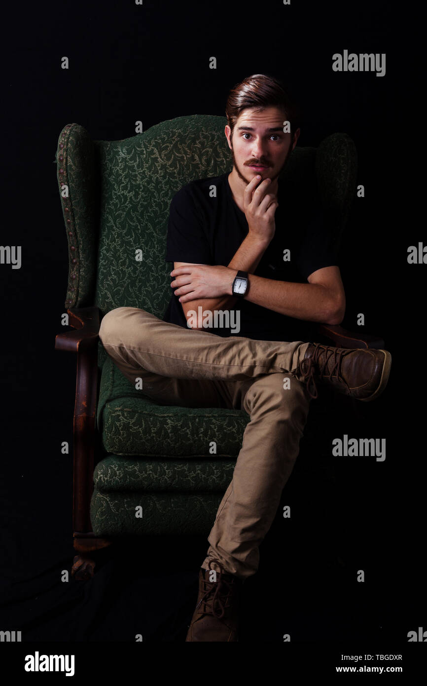 Young adult male sitting in vintage wingback chair with hand on chin, sinister or conflict expression. Color dark tones for dramatic effect, dark and  Stock Photo