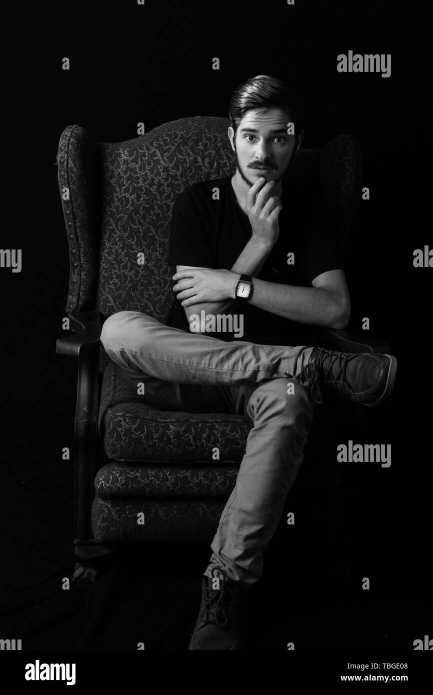 Young adult male sitting in vintage wingback chair with hand on chin, surprise expression. Monotone, black and white for dramatic effect. Concept for  Stock Photo