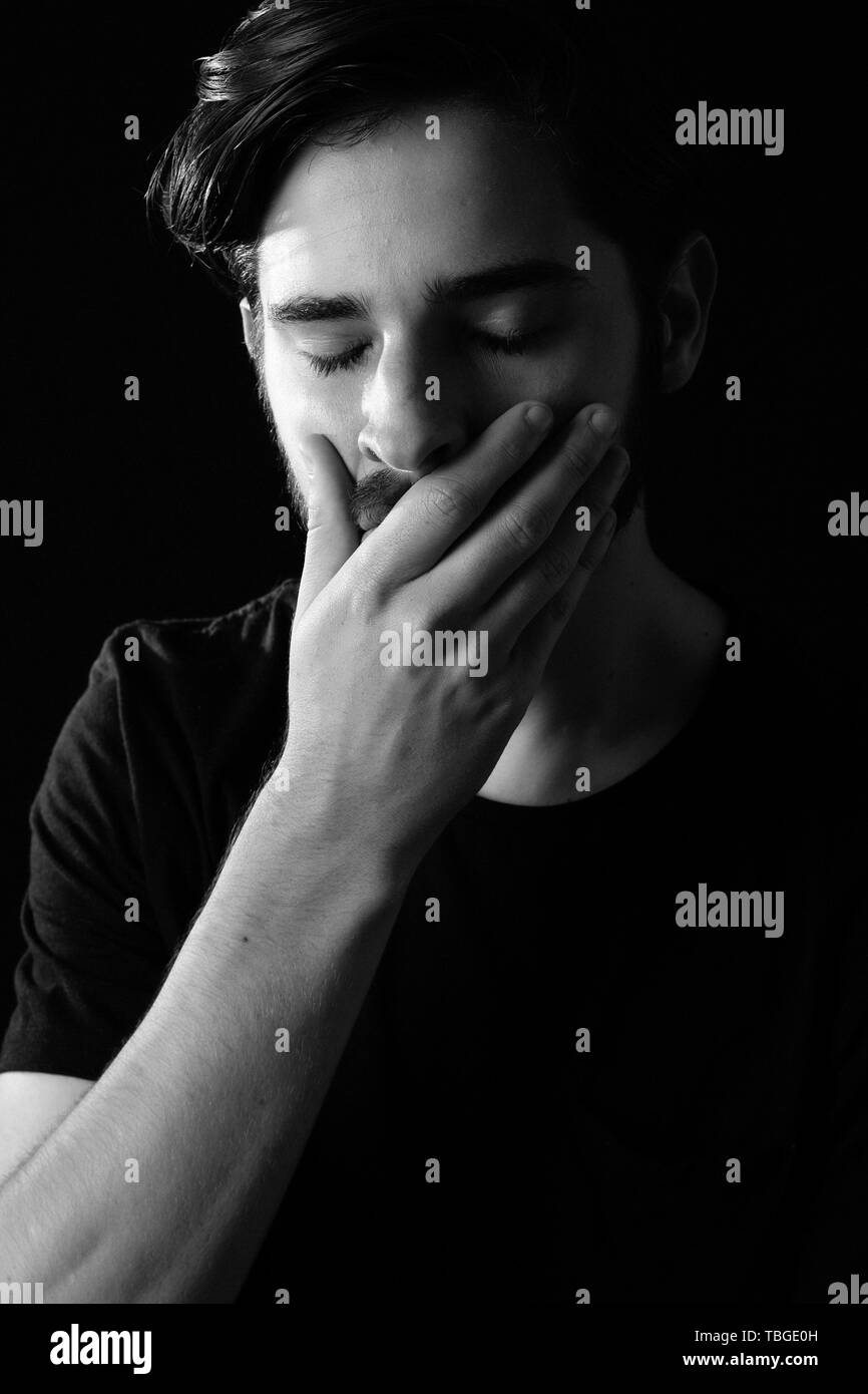 Close up of young adult male eyes closed hand over mouth in shock or horror. Monotone, black and white for dramatic effect, dark and moody series. Con Stock Photo