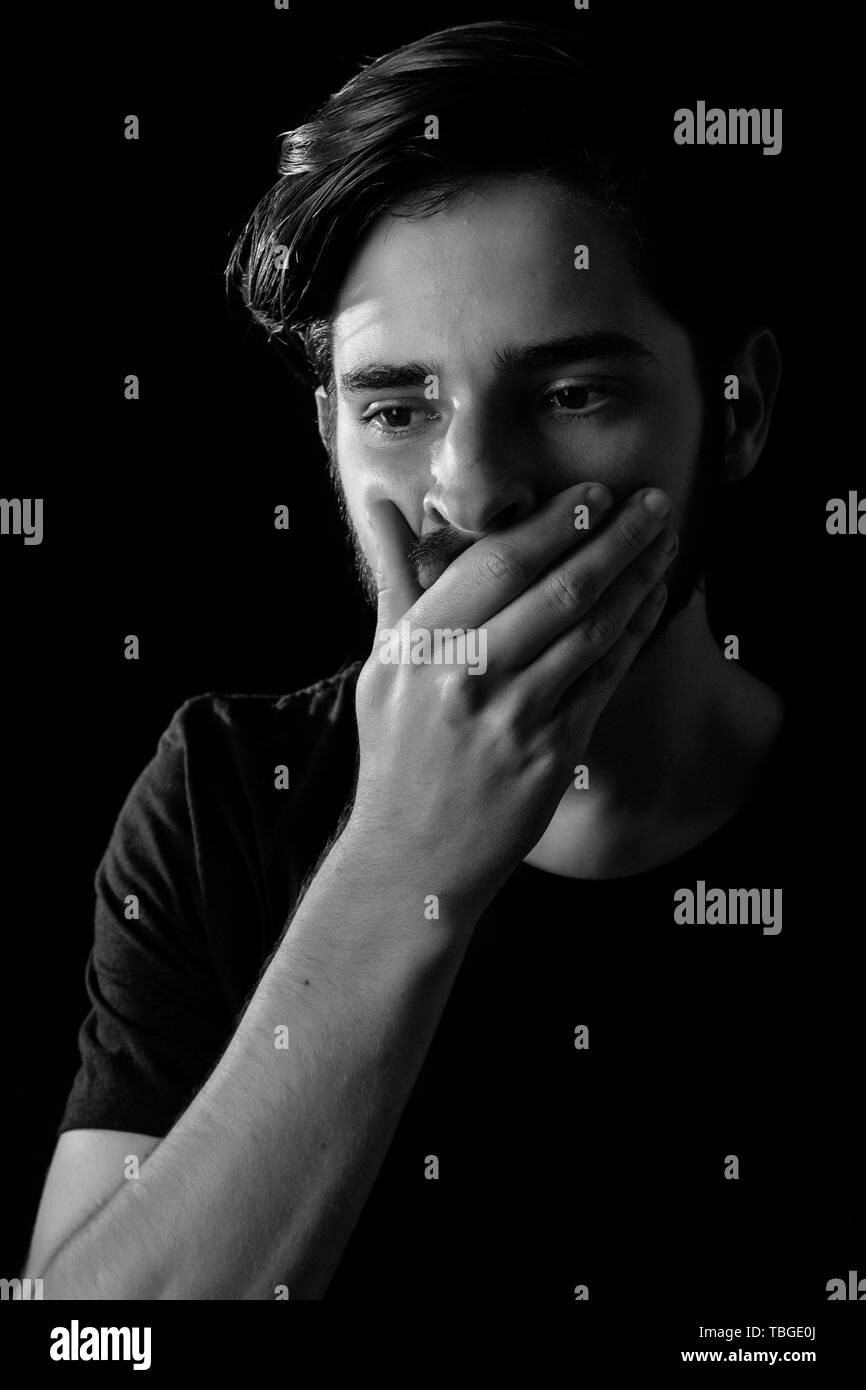 Close up of young adult male eyes open hand over mouth in shock or horror. Monotone, black and white for dramatic effect, dark and moody series. Conce Stock Photo