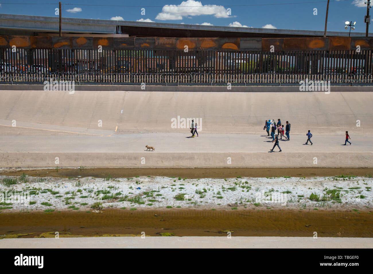 A group of migrants, including minors, walk along the banks of the Rio Grande towards a gap in the Border Wall under a new highway overpass near downt Stock Photo