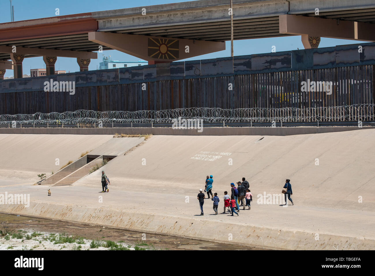 A group of migrants, including minors, walk along the border wall, past concertina wire strung along the banks of the Rio Grande near downtown El Paso Stock Photo