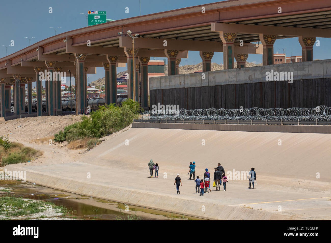 A group of migrants walk along the banks of the Rio Grande towards a gap in the Border Wall under a new highway overpass near downtown El Paso, Texas, Stock Photo