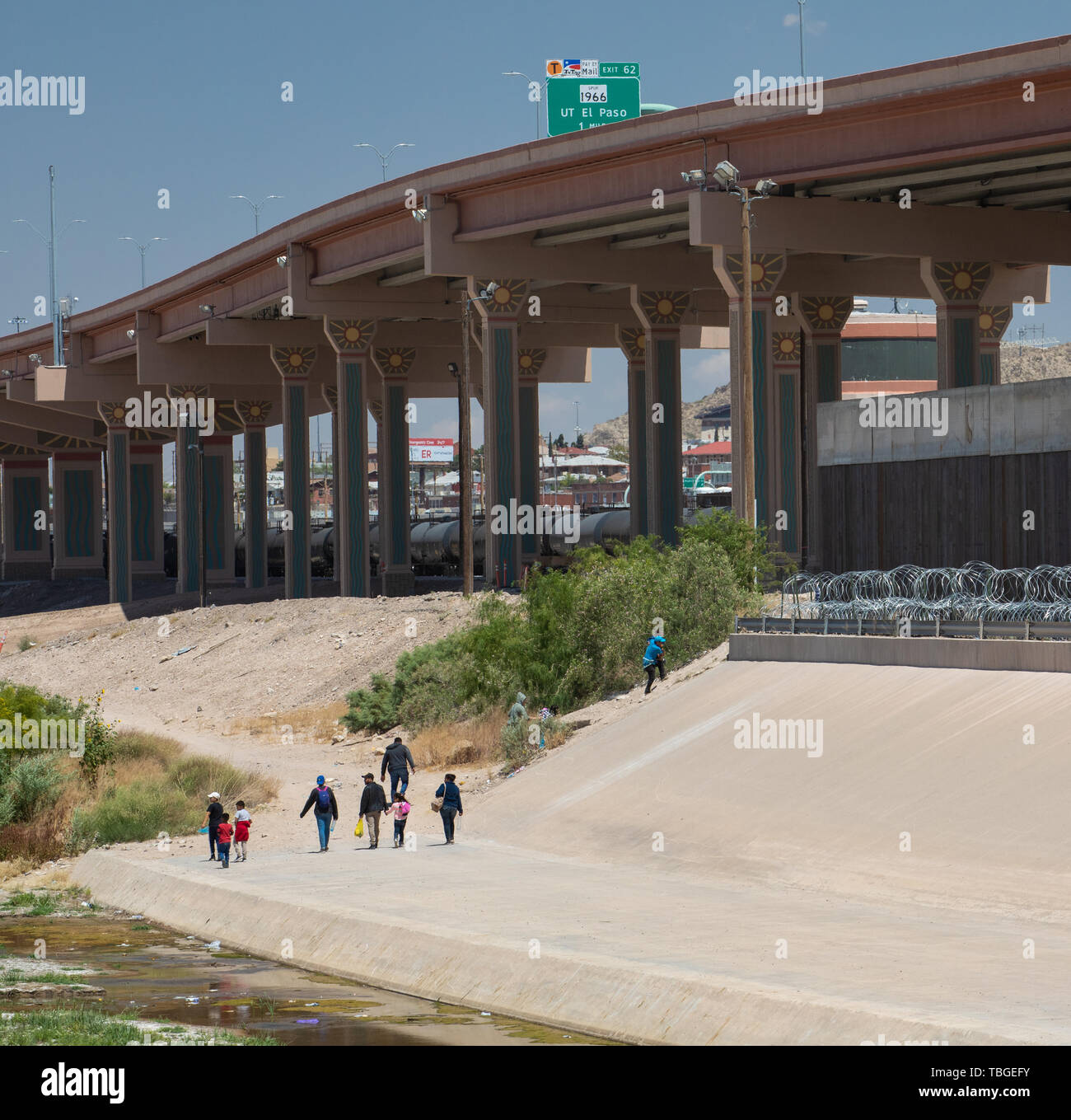 A group of migrants walk along the banks of the Rio Grande towards a gap in the Border Wall under a new highway overpass near downtown El Paso, Texas, Stock Photo