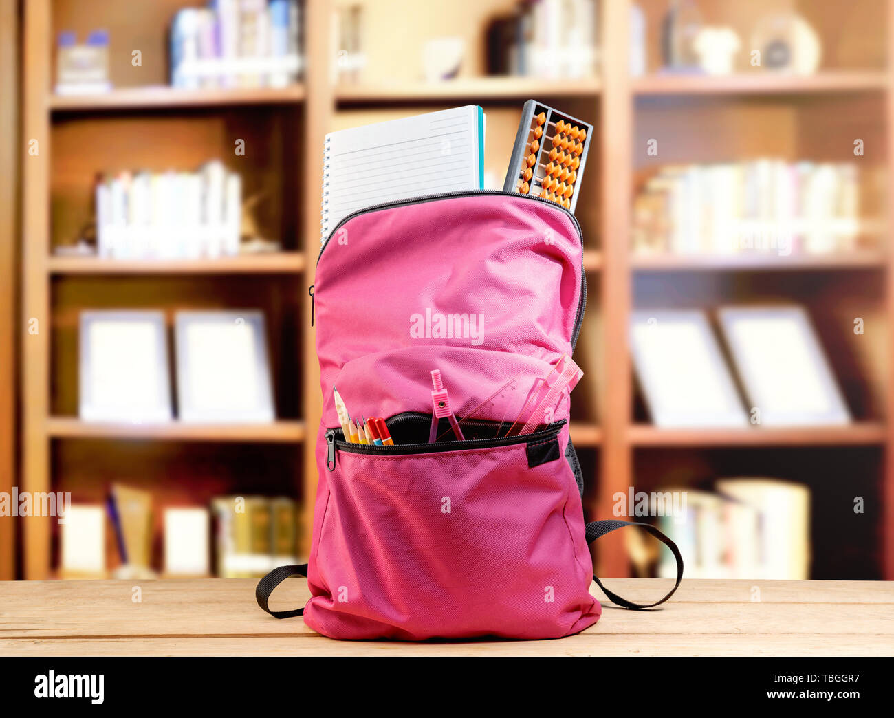 Pink backpack with book and different stationery on the wooden table in library. Back to School concept Stock Photo