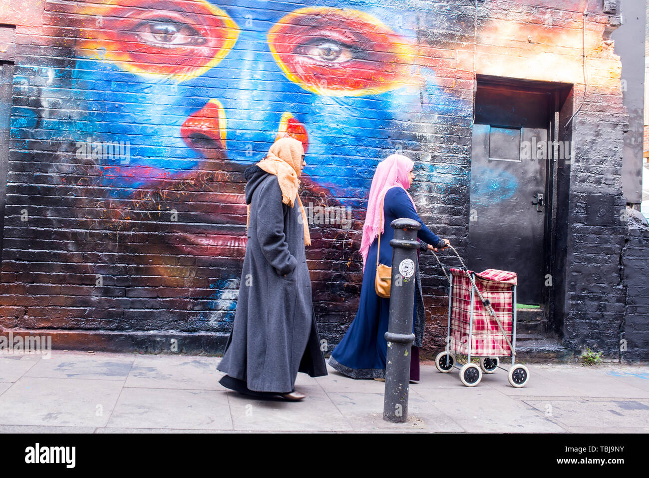 Brick Lane, Shoreditch, London, England, UK - April 2019: Two Indian women wearing hijabs walking in Brick lane next to a wall covered in graffiti mur Stock Photo