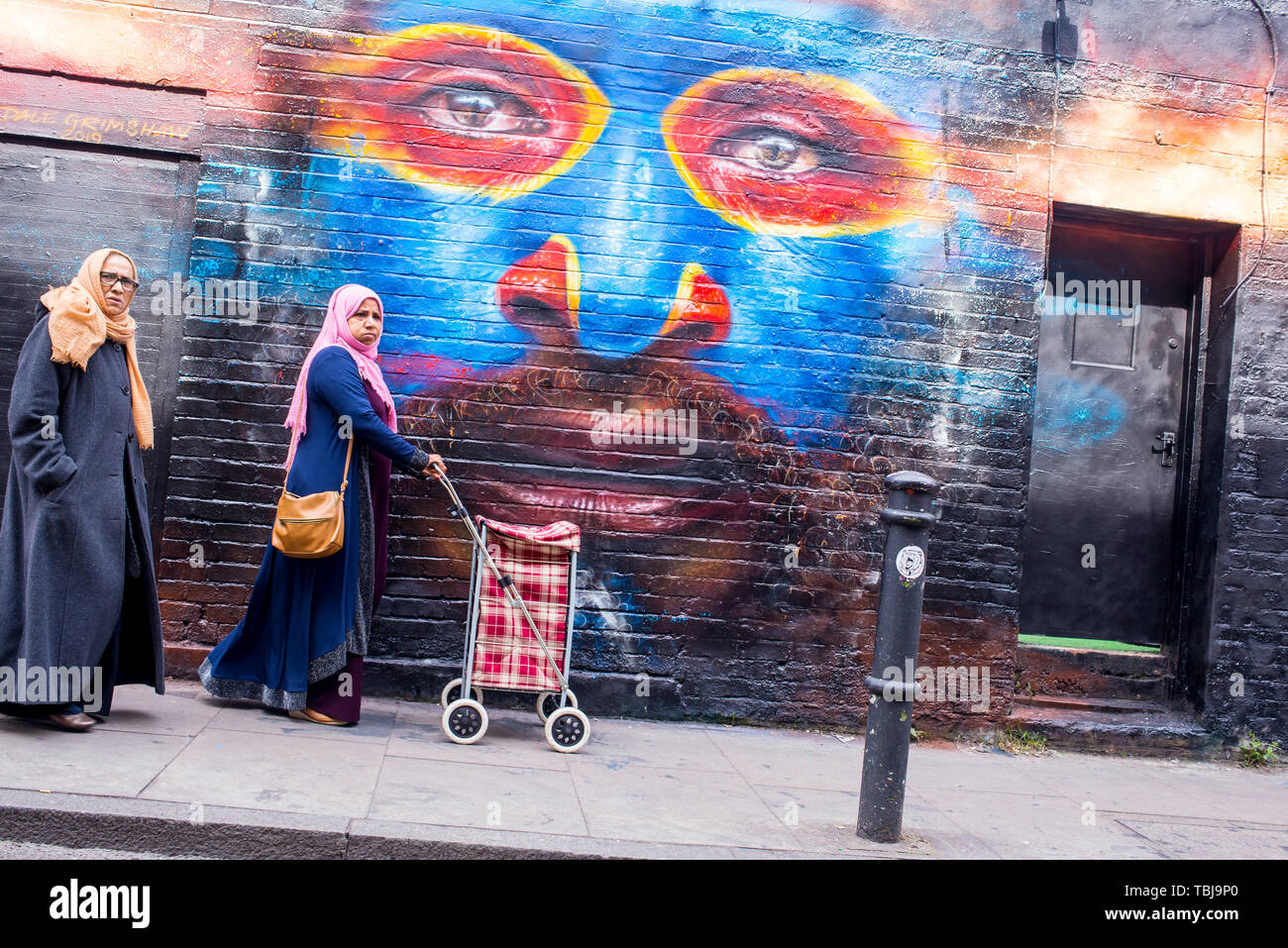 Brick Lane, Shoreditch, London, England, UK - April 2019: Two Indian women wearing hijabs walking in Brick lane next to a wall covered in graffiti mur Stock Photo
