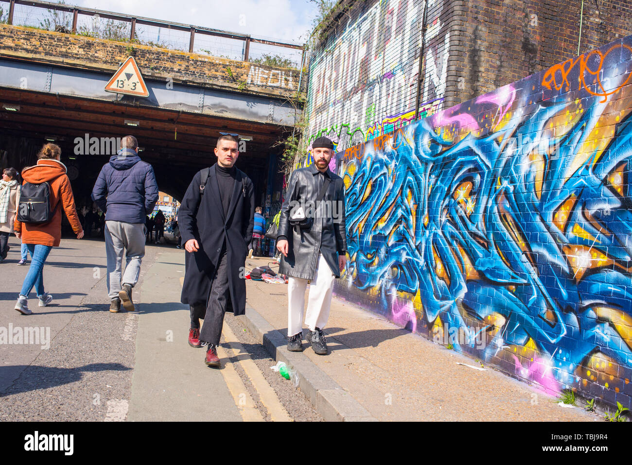 Shoreditch, London, England, UK - April 2019: Two hipster men walking in Wheler street next to a wall covered in graffiti mural street art in near Sho Stock Photo