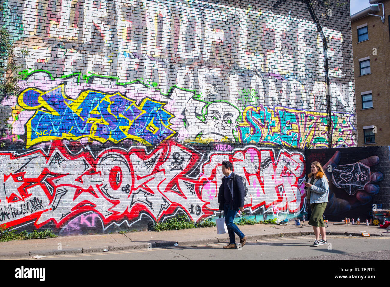 Shoreditch, London, England, UK - April 2019: People walking in Wheler street next to a wall covered in graffiti mural street art Stock Photo