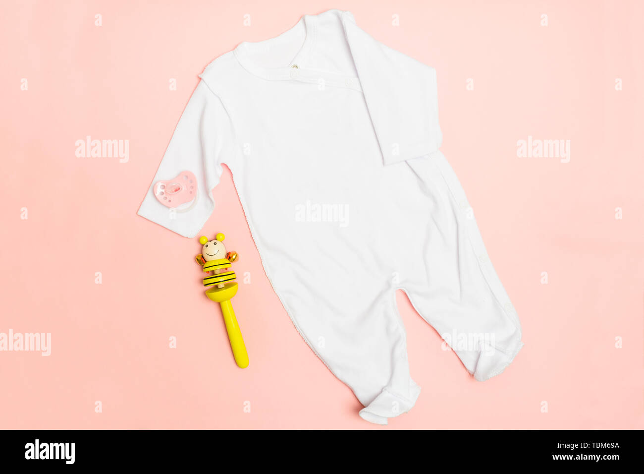 White textile bodysuit with yellow wooden rettle for newborn baby on pink background. Copy space. Flat lay. Top view Stock Photo