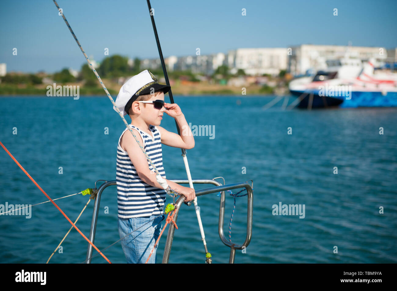 happy little boy in white captain hat and striped tank top standing on yacht board adjusting sunglasses in sea port during summer travel Stock Photo
