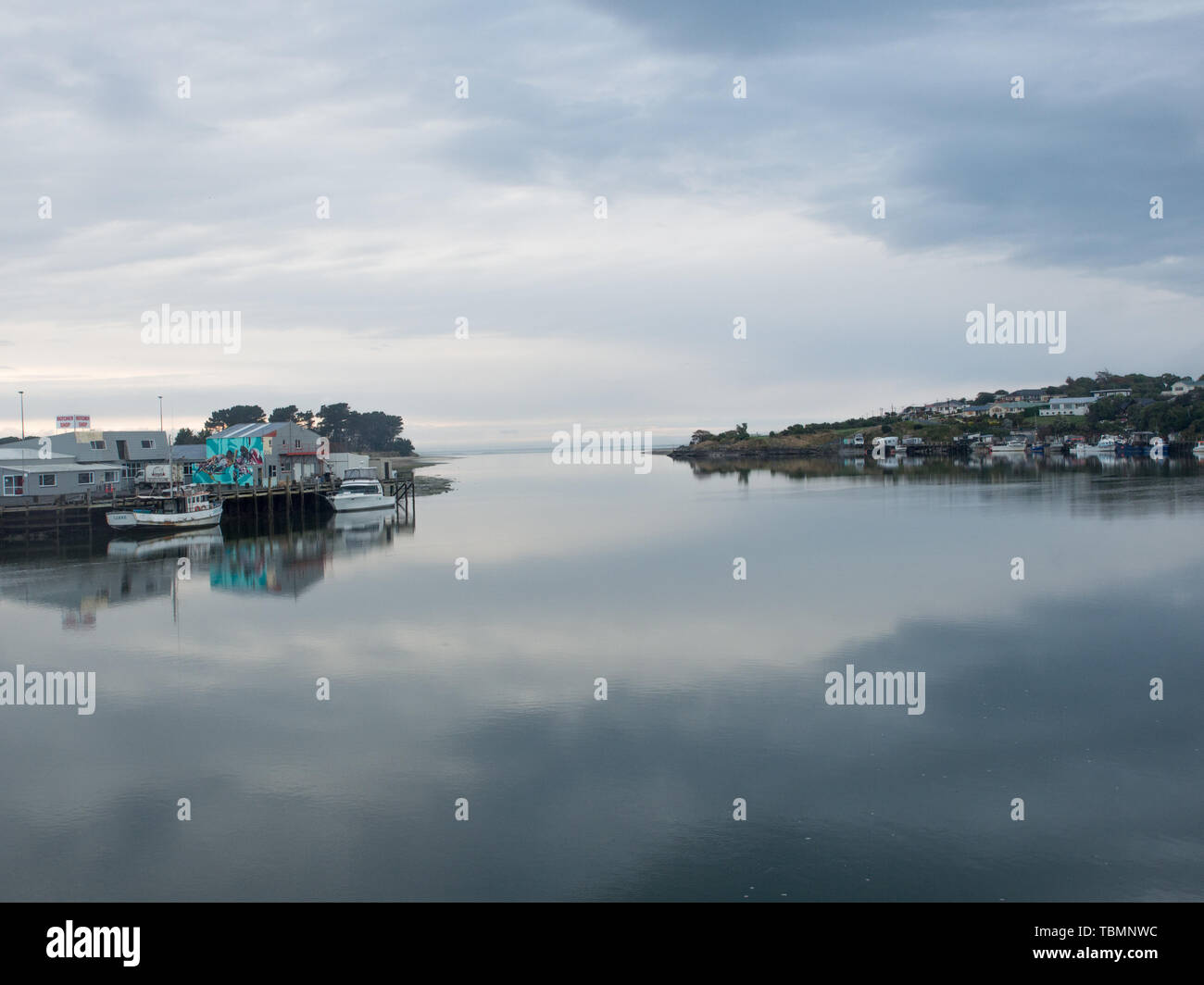 Fishing boats by the wharf, houses on the hill, gray sky above, calm water below, Jacob River estuary, Riverton Aparima, Southland, New Zealand Stock Photo
