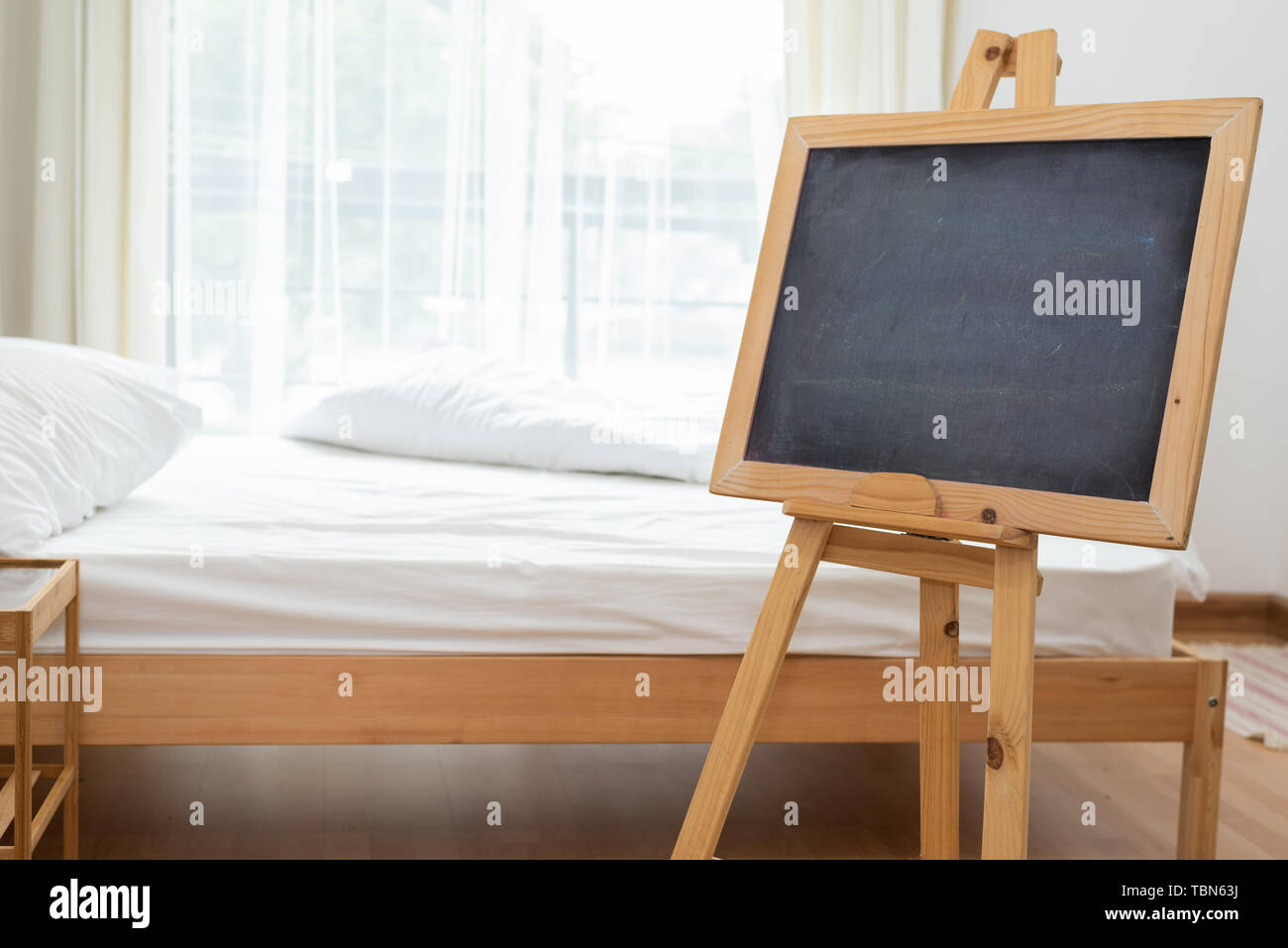 Black chalkboard on easel stand in bedroom with white soft bed background. Interior and decoration concept. Stock Photo