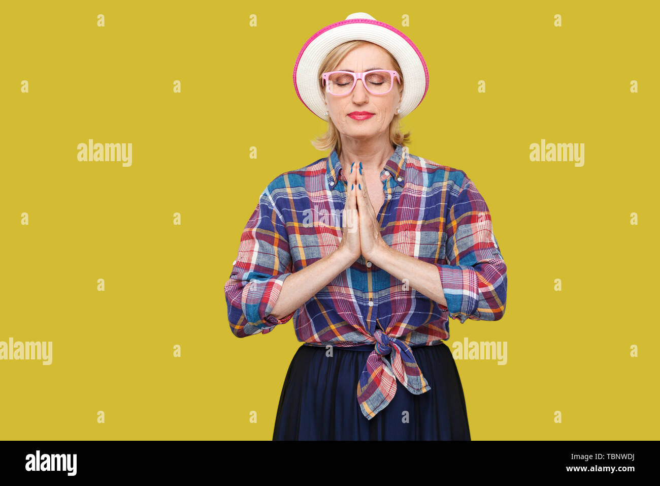 Portrait of calm modern stylish mature woman in casual style with hat and eyeglasses standing with palm hands, closed eyes, doing yoga pose exercises. Stock Photo