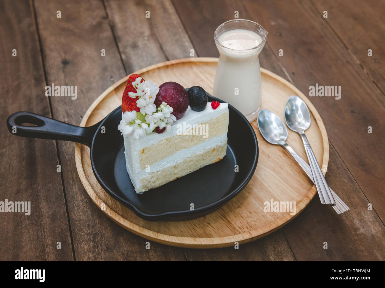 Sweet fruit cream cake cut serve with milk glass set on wooden plate with indoor low lighting. Stock Photo