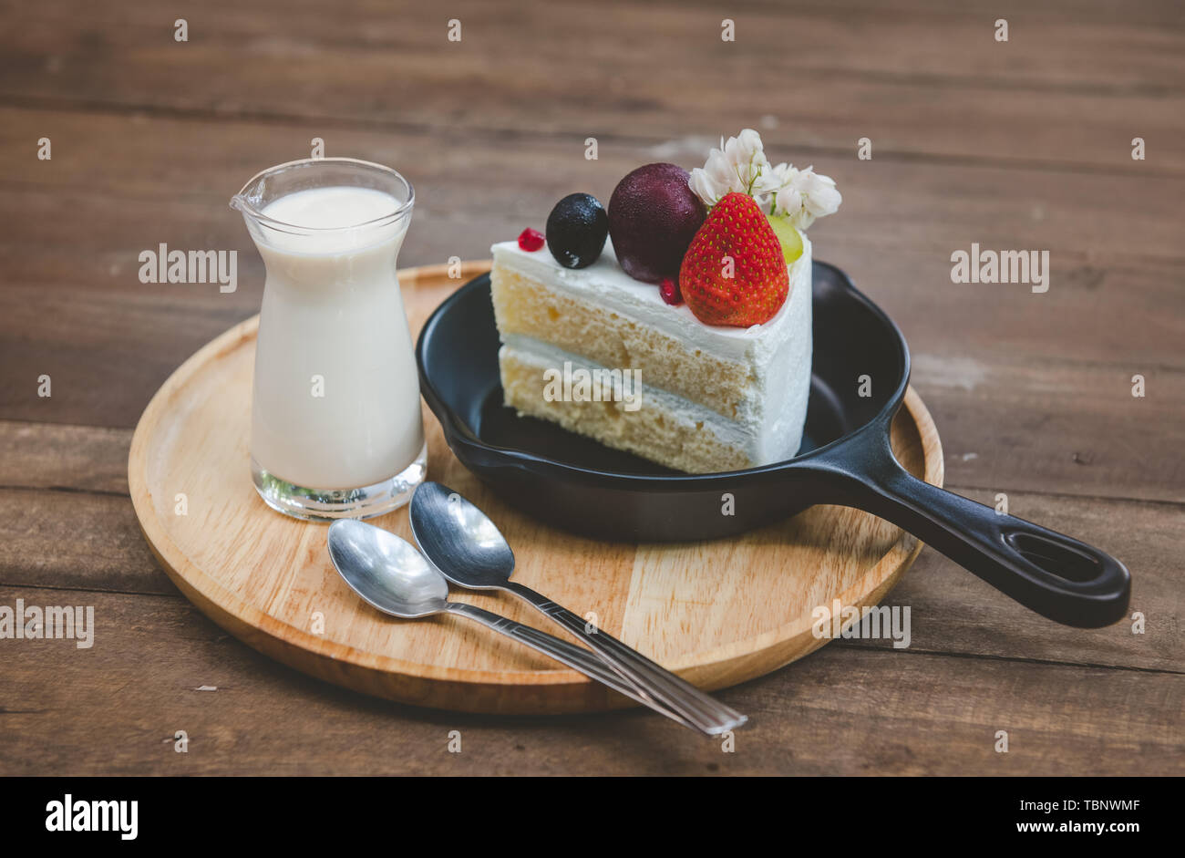 Sweet fruit cream cake cut serve with milk glass set on wooden plate with indoor low lighting. Stock Photo